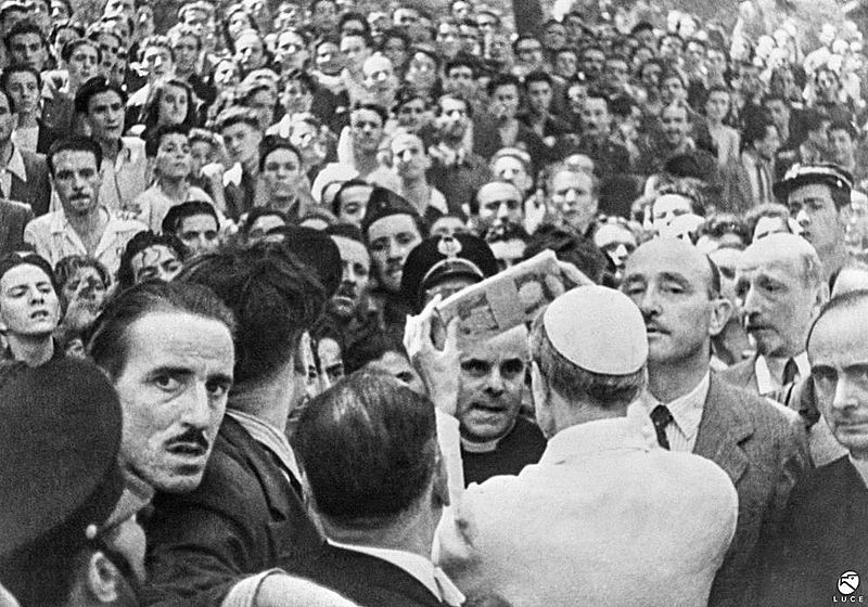 Papa Pio Xii distribuisce aiuti in denaro dopo i bombardamenti di Roma a Piazza San Giovanni, 13 agosto 1943. Nell'angolo di destra è riconoscibile il Sostituto segretario di Stato Giovanni Battista Montini, futuro Paolo VI